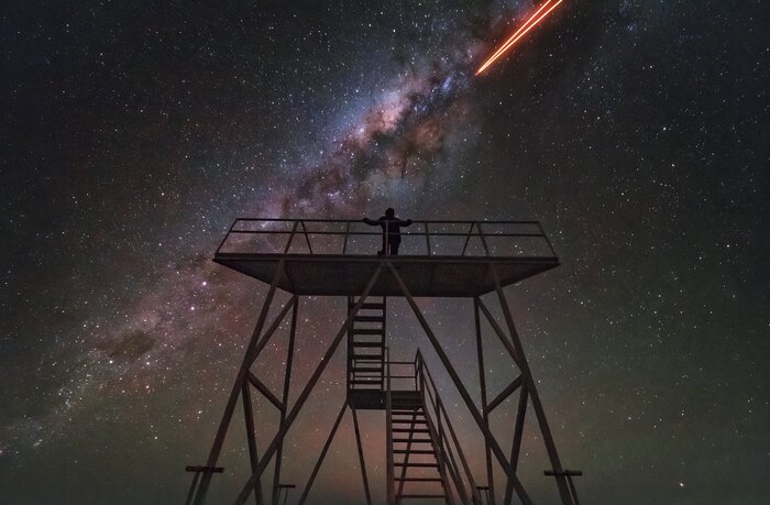 At the centre of the image is the silhouette of a person standing on a tall viewing platform, at least 5 metres high and almost just as wide. It has a two-level staircase leading up to it. The dark foreground silhouette of stairs and platform contrasts with the night sky filled with stars, showing blue and green hues, and the cloudy, white Milky Way plane reaching across, diagonally. At the top right, two orange laser beams seem to come out of the galactic plane, going diagonally to the top of the image.