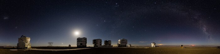 The image is an elongated horizontal panorama. It shows eight telescope domes in the bottom fifth of the image. The moon lights the telescopes and the desert from behind, creating a haze across, but above it the sky is pretty dark and full of stars. In the right half of the image is our Milky Way galaxy –– an arc made dense by white points and hues of pink and purple.