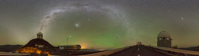 This is a very wide panoramic image of the night sky from an observatory. The dark sky is dominated by a green glow, with some white smudges showing, and the white and purple Milky Way arches across the sky. Three telescopes are located at different positions: a big, rounded, white building to the right, a tiny grey one to its left, and a smaller white one on a brown rocky hill even further to the left.