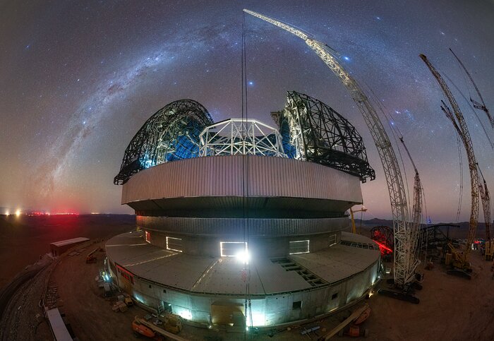 A night image of a partially-built telescope under the stars. The image uses a distorted projection, which creates a focal point in the centre which is closer to the viewer, and which everything bends around. The middle of the picture is taken up by the dome building of the ELT, with a crane on the right that extends from the bottom right of the image, curving up to the middle of the image at the top. Its tether line splits the image in half horizontally. The dome of the ELT is open, showing the beams that hold the main structure in place, around it, are the beams that will make the doors once panelling is attached to it. Above the ELT on the left is the Milky Way, extending from the horizon about a third of the way up the image, meeting the top of the crane in top middle of the image. The Milky Way looks like a blue and purple wispy cloud, with points of white, the brighter stars.