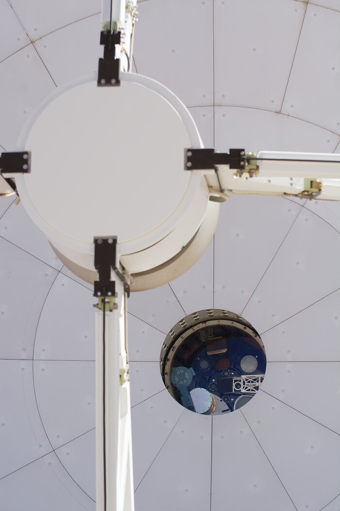 The image is a close up of one the ALMA antennas. In the centre left foreground of the image, is a circular structure held by four support beams that continue outside of the boundaries of the image. In the background is the grey dish made up screwed on panels. To the bottom right in the background is a hole for the receiver cabin, filled with circular and rectangular instruments on a blue plate.