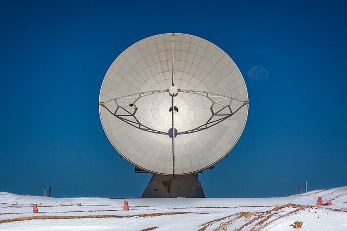 An enormous white dish stands on its side, as if looking out onto the horizon behind the photographer who took this picture. It’s held up by a large stand, and four beams come from the top, bottom and sides of the dish to meet in the middle of the huge antenna. Nothing else is visible around this radio telescope, it dominates the landscape of the image, and the sky behind it is completely free of clouds. Just to the right of the dish, the moon is faintly visible, looming over the telescope’s shoulder.