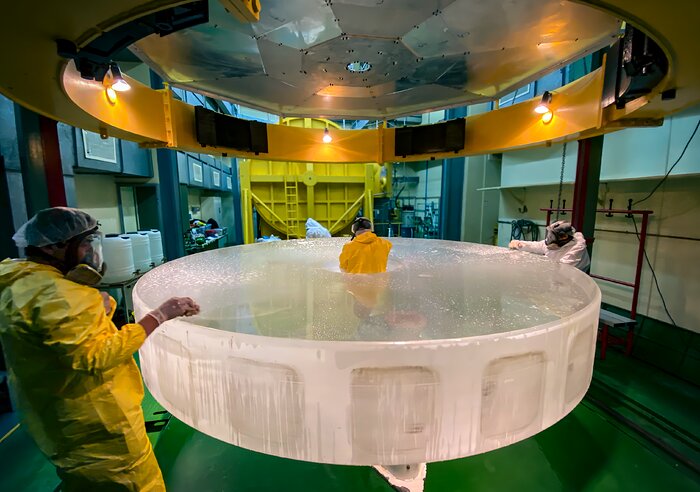In the centre of the image lies a white cylinder block with engineers and technicians standing in the centre, their torso peeking out of the hole. They are standing around the block wearing bright hazmat suits, face shields, clear gloves, and hairnets. They are cleaning the surface layer of the mirror blank.