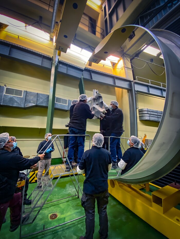 Two people are standing on a ladder to reach up and fix the last piece over the whole in the centre of the mirror. There are no imperfections or warps in the mirror, making it appear as though there are four people all holding up the centre piece. Team members are standing to the left of the ladder, wearing face masks and hair nets.
