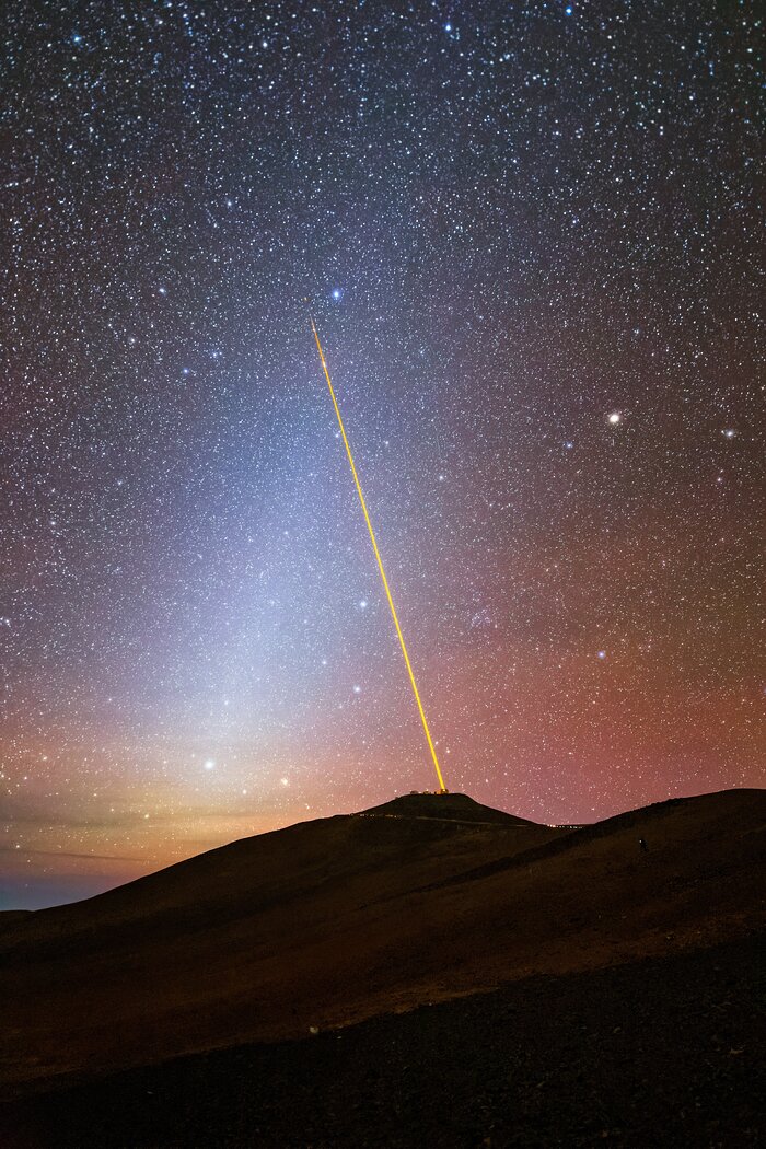 In the centre of the image is a bright, thin yellow line extending from a point on a black mountain that fills the bottom third of the image. The yellow laser extends from the centre right of the image at the top of the mountain to two-thirds of the way up the image on the left-hand side. The yellow laser meets with a blurry streak of white that extends from just above the mountain on the left-hand side to the right of the image, forming an upside-down V. The sky just above the mountain is orange and pinkish, with thousands of bright white stars speckling the night sky.