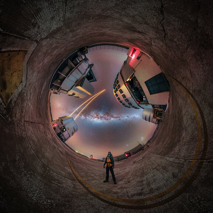 The image shows the Very Large Telescope (VLT) site at Cerro Paranal, captured as a 360 panorama. In the bottom centre you see one of the photographers looking up the sky, surrounded by the huge telescopes of the VLT. One of the telescopes is shooting lasers skywards. The Milky Way band shines bright upon the site and the VLT telescopes seemingly tower over the photographer, appearing miniscule in comparison.