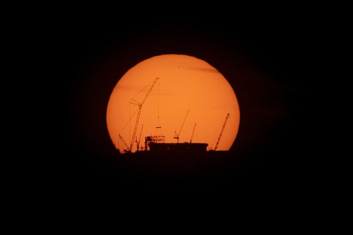The base of a cylindrical building and some huge cranes are silhouetted against the orange disc of the Sun.
