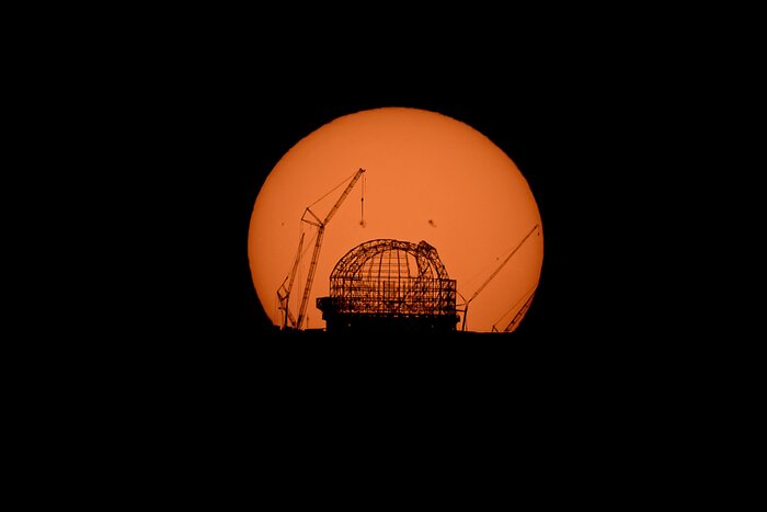 The mesh skeleton of a huge dome and cranes are silhouetted against the orange disc of the Sun.