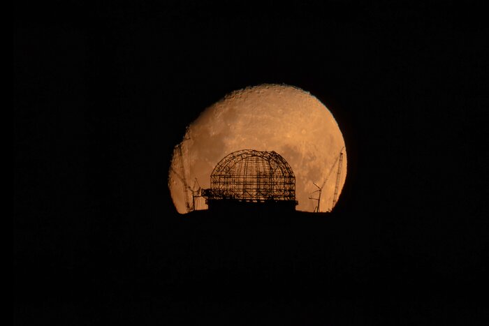 The mesh skeleton of a huge dome and cranes are silhouetted against the Moon.