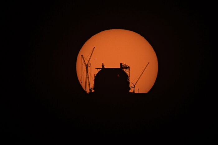 A dark huge dome and some cranes are silhouetted against the orange disc of the Sun.