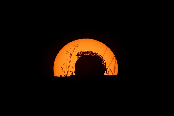 A dark huge dome and some cranes are silhouetted against the orange disc of the Sun.