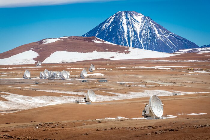 The image shows a partly snowy desert, on which some white antennas are distributed, with some gathered together in the middle-left part of the image. In the background a dark mountain arises in front of the blue sky, also with snow on it.