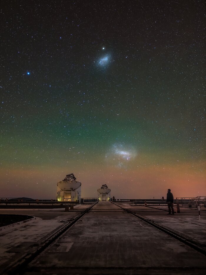 The image shows two of ESO’s small Auxiliary Telescopes facing the vastness of the two Magellanic Clouds on the sky. The interplay between those two pairs and the human silhouette on the right side of the picture, displays the immensity of the cosmos compared to the short-lived time of humanity on Earth.