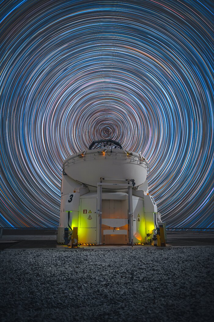 An open white dome, with part of a telescope peeking through its opening at the top. The sky in the background is full of countless concentric white-ish circles, centered at the top of the telescope.