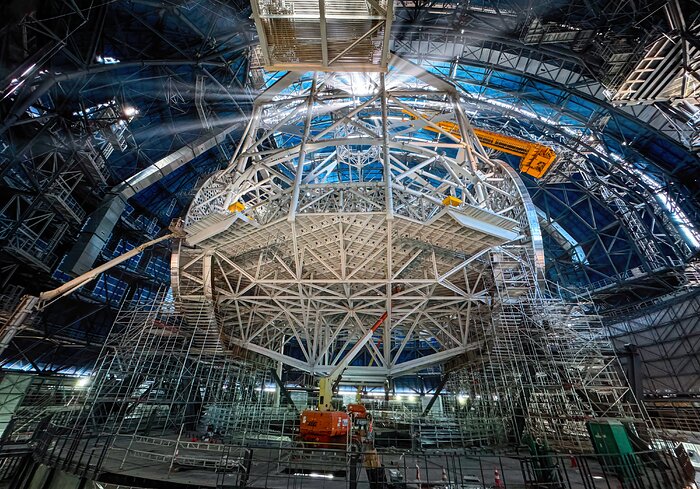 This image shows the interior of the Extremely Large Telescope's (ELT) dome. The telescope structure, supported by struts, takes up most of the image in the centre, with some cranes around working. The gigantic doors of the dome are open, allowing sunlight to shine through.