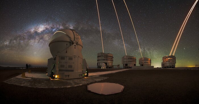 The panoramic image shows one of the Auxiliary Telescope in front with a closed dome. In the background the four Unit Telescopes are observing the night sky while their lasers light up the image in a warm orange light. The sky above Paranal is so pristine that the entire Milky Way band is visible on the sky with a particularly bright patch in the middle. Additionally, the horizon is lit up by green, yellow shimmer caused by the airglow, the natural glow of the atmosphere only visible in the darkest places.