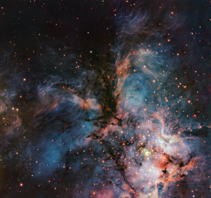 A telescope image of a cosmic gas cloud. The cloud is full of wispy filaments that shine brightly in shades of pink and orange, especially in the lower-right part of the image, where there is a higher concentration of stars. Some dark dust lanes partially block some areas of the nebula.