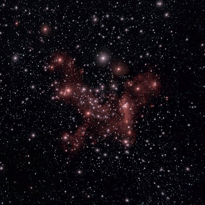 This image has a black background in which some thousands of light sources are visible. Some of these stars are closer and some are further away, looking like simple white dots on the night sky. In the centre of the image, more closer stars are gathered. These centred stars are embedded in a pinkish haze of gas.