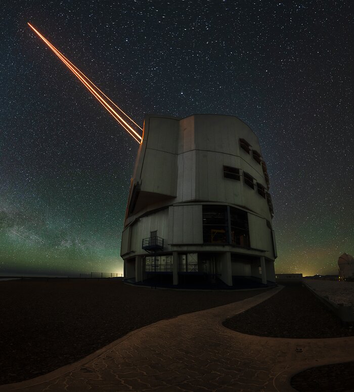 The image shows a large cylindrical, metallic dome, from which four laser beams emerge on the left. These orange beams seem to pierce through the sky. In the background the night sky emerges with its thousands of stars, underlining the majestic nature of the telescope.