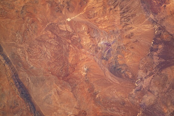 This image shows an aerial view of a wide rocky landscape. The ground is various shades of reds and greys, marked by contours. Just below the centre of the frame is a black dot with a grey line circling around it then leading straight down. This stands out only slightly compared to the rest of the features in the image.