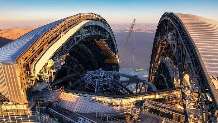 This aerial image shows a huge dome from above, partially split into two halves. Along the edges of each half there’s a complex network of metallic beams and catwalks. The opening reveals part of the metallic frame of a huge telescope inside the dome.