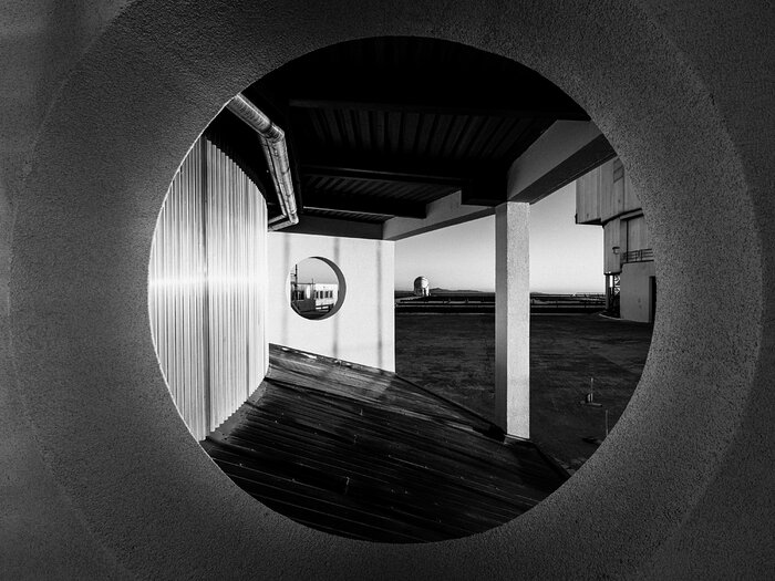 A black and white image showing a small, white telescope dome framed by a circular hole in a concrete wall in the foreground.