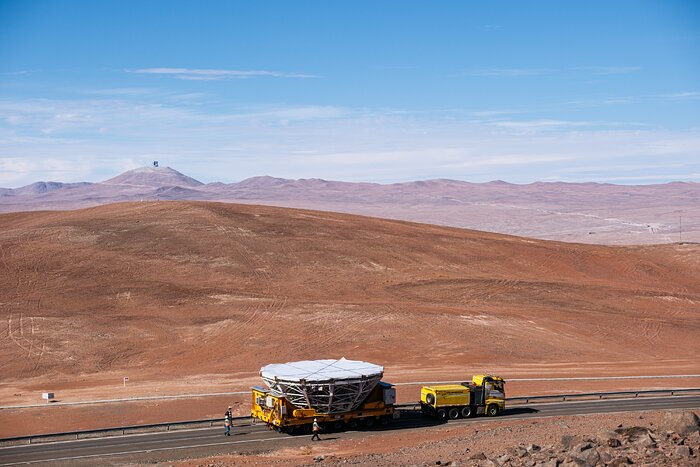 A desert takes up most of the frame in this image. Close to the camera, a yellow truck can be seen carrying a telescope mirror and its support frame, and, in the background, a small grey sphere (the ELT) appears against the blue and cloudy sky.