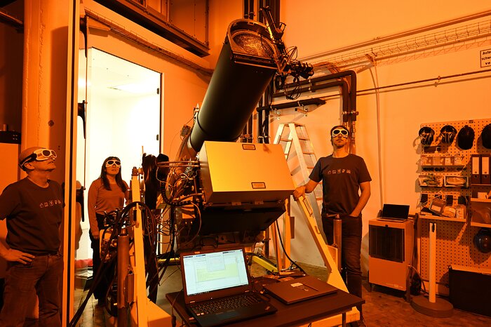 Three people in a lab, wearing safety goggles. They’re next to a large black tube resembling a telescope. The entire room is bathed in orange shades, cast by light emitted by the tube.