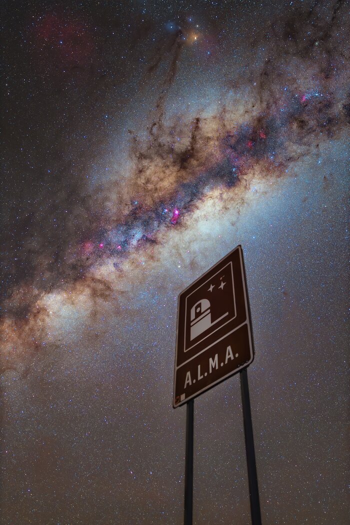 The image shows the ALMA road sign, with the night sky in the background. The top half of the background features the Milky Way, going diagonally, showing darker and purple tones at its centre, and orange and brown colours on its ends. In the rest of the background, a lot of stars, bright white spots in the sky, can be seen.