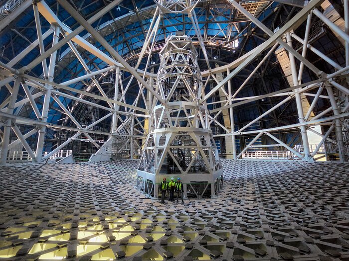 The image shows the interior of the Extremely Large Telescope. The metallic grey main structure can be seen in the entire picture, with three people standing in front of it, in hard hats and green safety vests. The people are much smaller than the telescope’s structure.