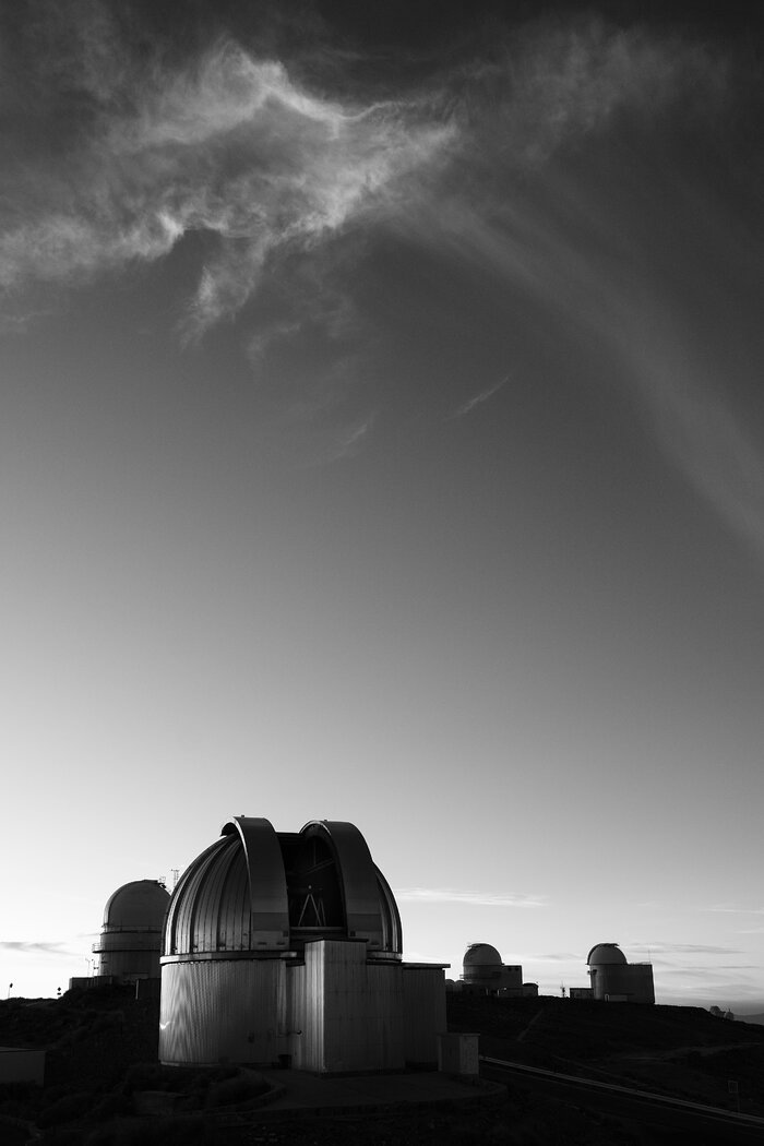 A black and white photograph in portrait. In the bottom third of the image, in the foreground, is a cylinder-shaped building with a shiny dome, The dome is split into two halves with an opening between, and a metal structure is visible in the dark gap. Three similar-shaped buildings are in the background, one to the left of the main building and two to the right, mostly in shadow. The ground is shades of dark grey to black. The sky is a gradient from light grey at the bottom of the image to dark grey at the top. The top third of the image is filled by a wispy white cloud.