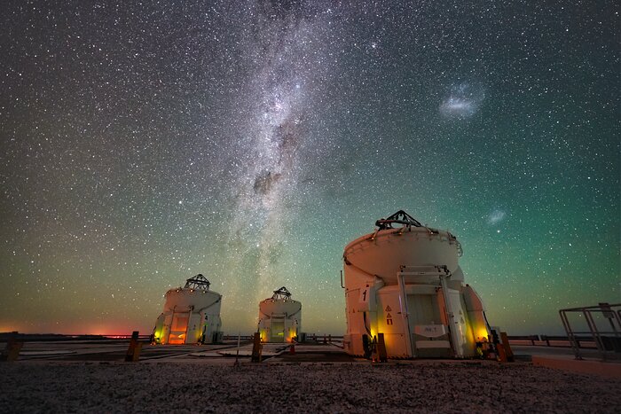 The image shows three white Auxiliary Telescopes, at the bottom of the frame. In the background, in blue and green colours, and yellow and red towards the horizon, the night sky is full of stars, with the white Milky Way band vertically in the middle of the picture. On its right, the two white Magellanic clouds can be seen.