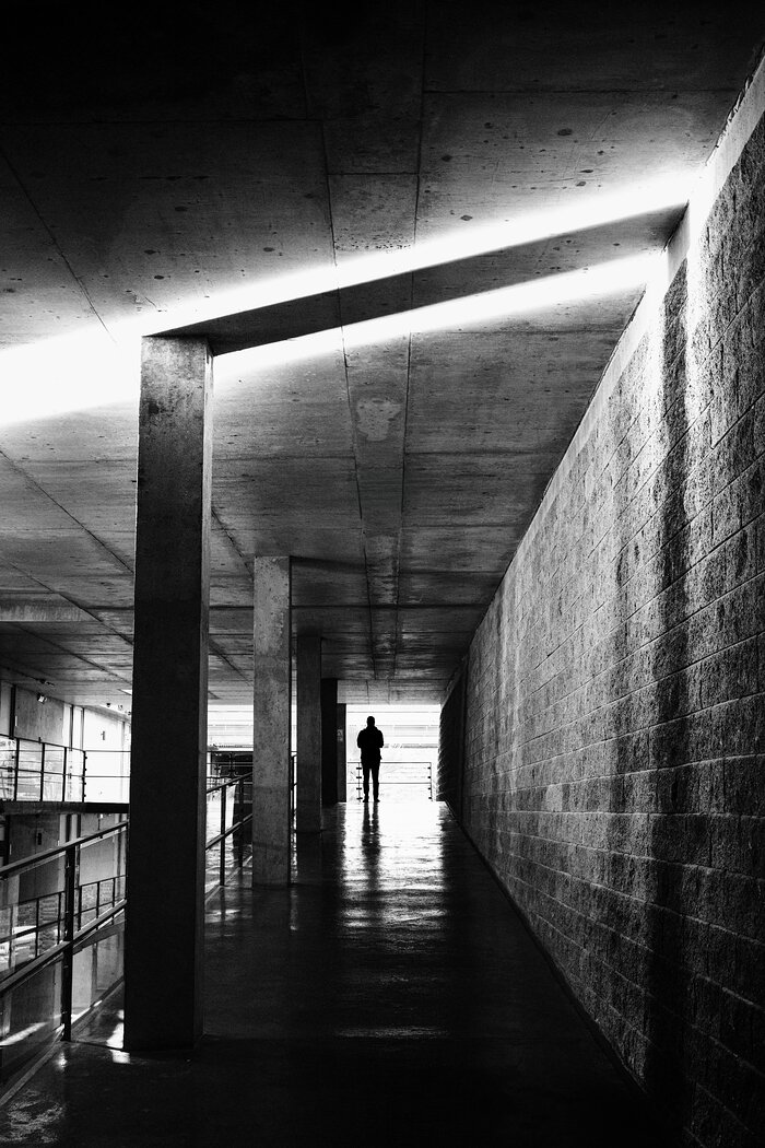 A vertical black-and-white photograph of a long concrete corridor inside the Paranal Residencia. A rough concrete wall extends along the right side, while a railing and a row of rectangular columns line the left. A bright diagonal strip of sunlight crosses the ceiling and is reflected on the dark floor. Near the distant, brightly lit end of the corridor, a small, silhouetted person stands at the top of the ramp. Strong straight lines converge around the figure, emphasising the depth of the corridor.