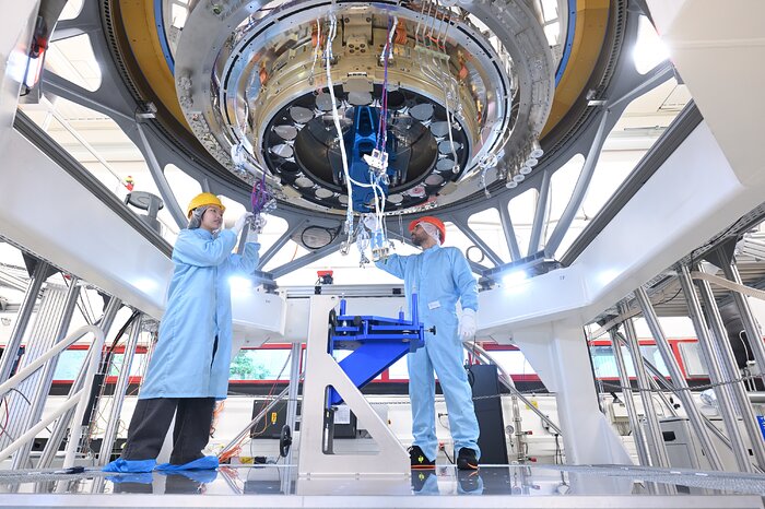 Two engineers in protective cleanroom clothing stand beneath the large circular centre wheel mechanism of the MICADO instrument. The wheel fills much of the upper part of the image, with cables, optical components and metal structures visible around it as the engineers inspect and work on the mechanism.