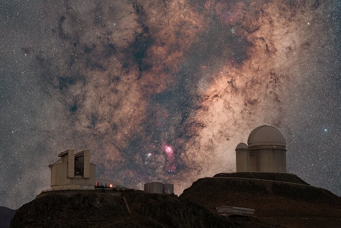 The image shows two white telescopes, on two hills on the bottom left and right of the frame. In the background, the night sky features a lot of stars, and, in the centre of it, a dark cloud, in tones of black and brown, that goes from top to bottom. In the bottom of this cloud, spots shining in red and pink, and some blue, appear. Another telescope can be seen further away in the picture.