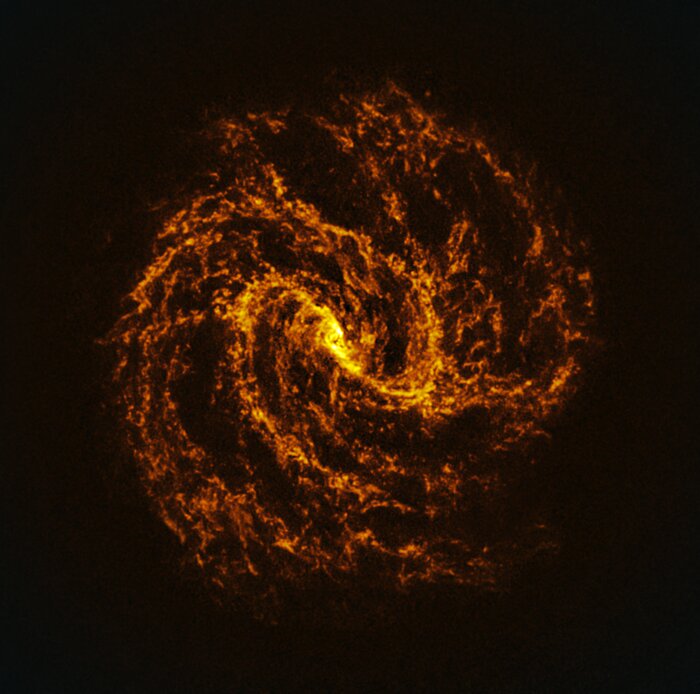This image shows an object made of two spiral arms connected at the centre, in shades of orange against a black background. The object is brightest at the centre and along the spiral arms. The spirals are composed of cloud-like threads that become more diffuse further away from the centre of the image, spreading out like loose tufts of wool.