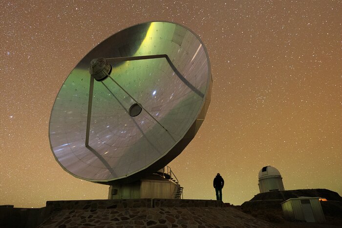 The image shows a reflective telescope dish, occupying a large portion of the frame. A person is standing close to the telescope, with their back to the camera, and wearing darker clothes. The sky in the background is filled with stars and orange to green undertones, and a white telescope, as well as a white container building, can be seen.