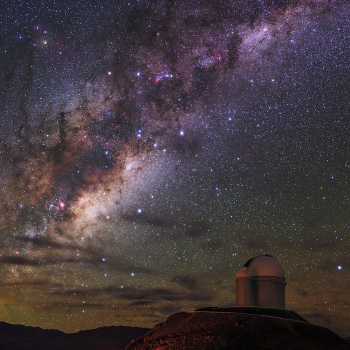 The radiant purple and white Milky Way dominates the star-speckled sky in this image, as it stretches from the top right of the frame to the bottom left. In the bottom right of the image, a telescope with its characteristic white dome examines the sky above from its perch at the top of a hill.